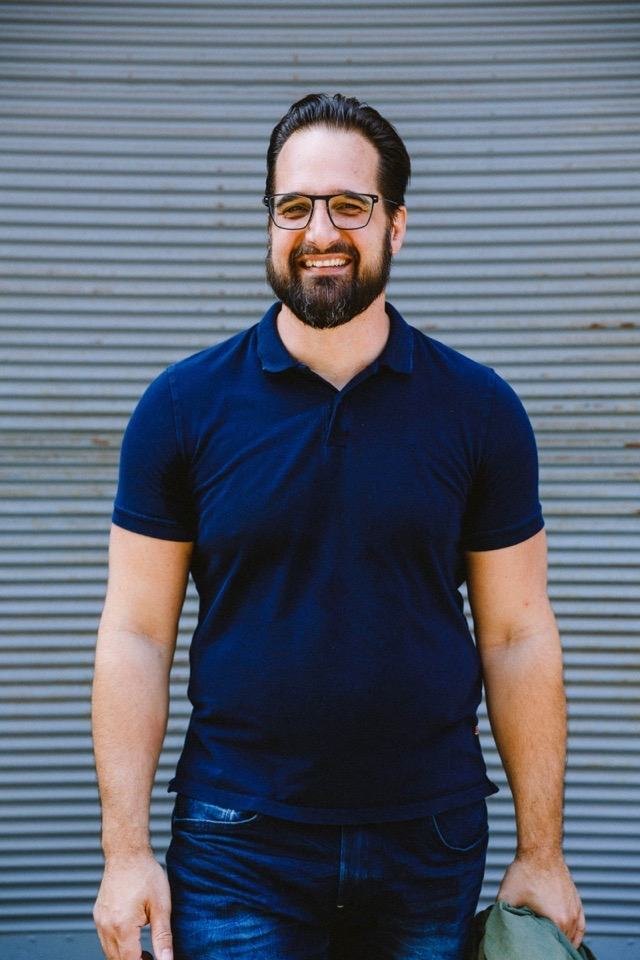 Mathias Ziehengraser mit Brille, die Jacke in der Hand, geht lachend auf die Kamera zu, hinter ihm eine graue Wellblechwand.
