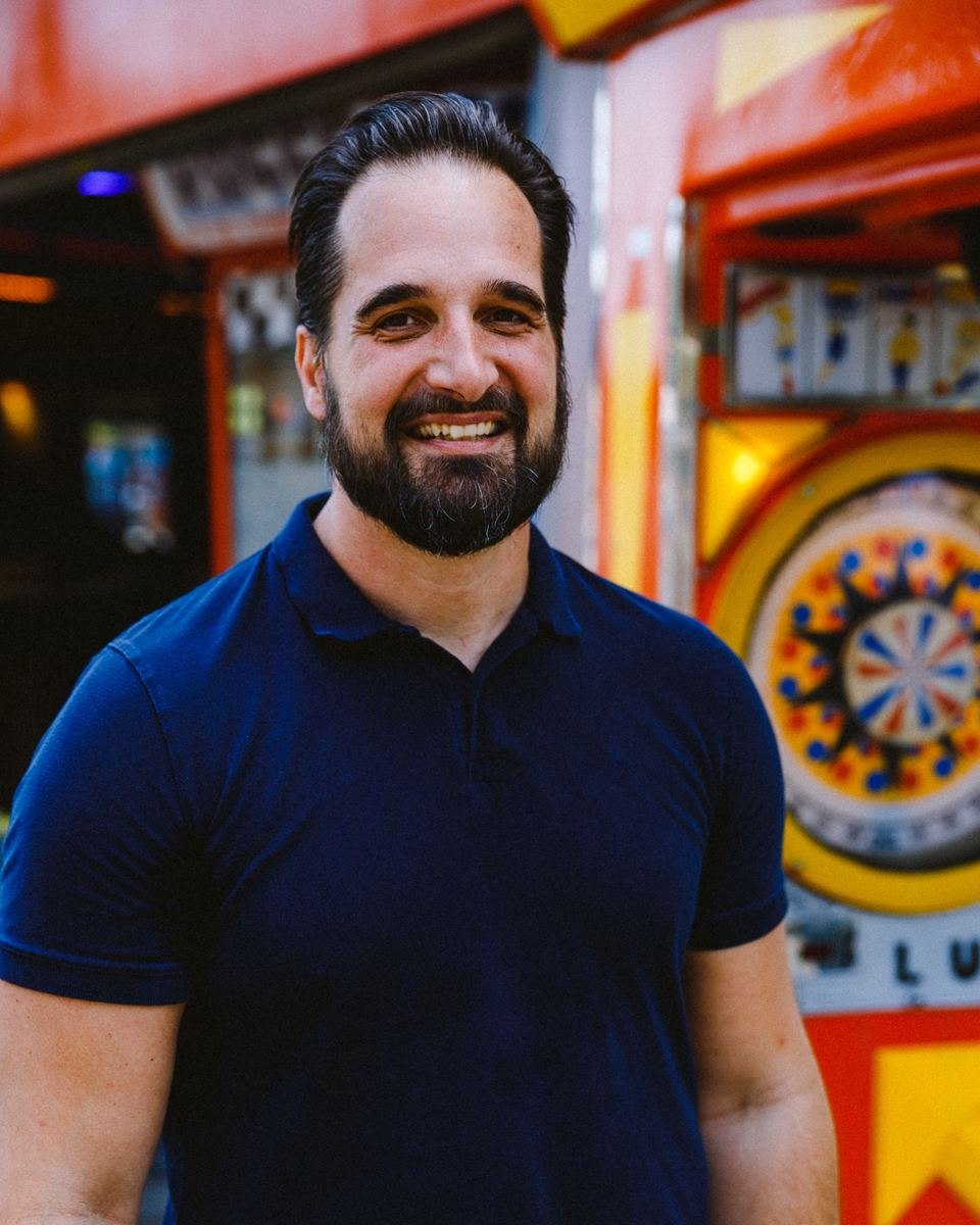 Mathias Ziehengraser lacht breit in die Kamera, hinter ihm ein bunt bemaltes Glücksrad an einer orangen Schaustellerbude.