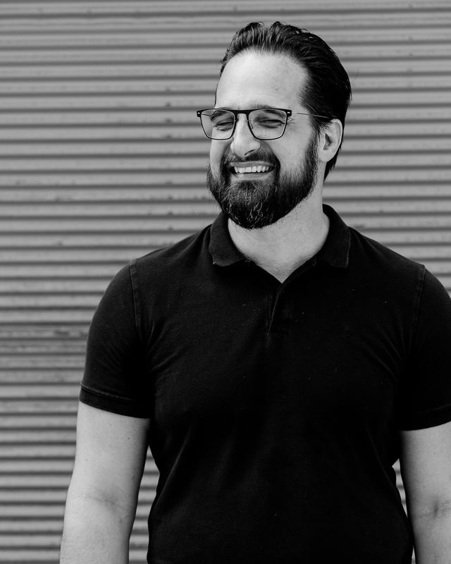 Schwarzweiß: Ich lache mit Brille und fast geschlossenen Augen zur Seite, hinter mir eine Wellblechwand.
